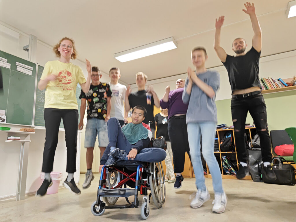 Ein Gruppenbild von TeamKnofti im Klassenraum. Ein Kind sitzt in der Mitte im Rollstuhl. Die anderen Schülerinnen und Schüler springen gleichzeit in die Luft und lachen.