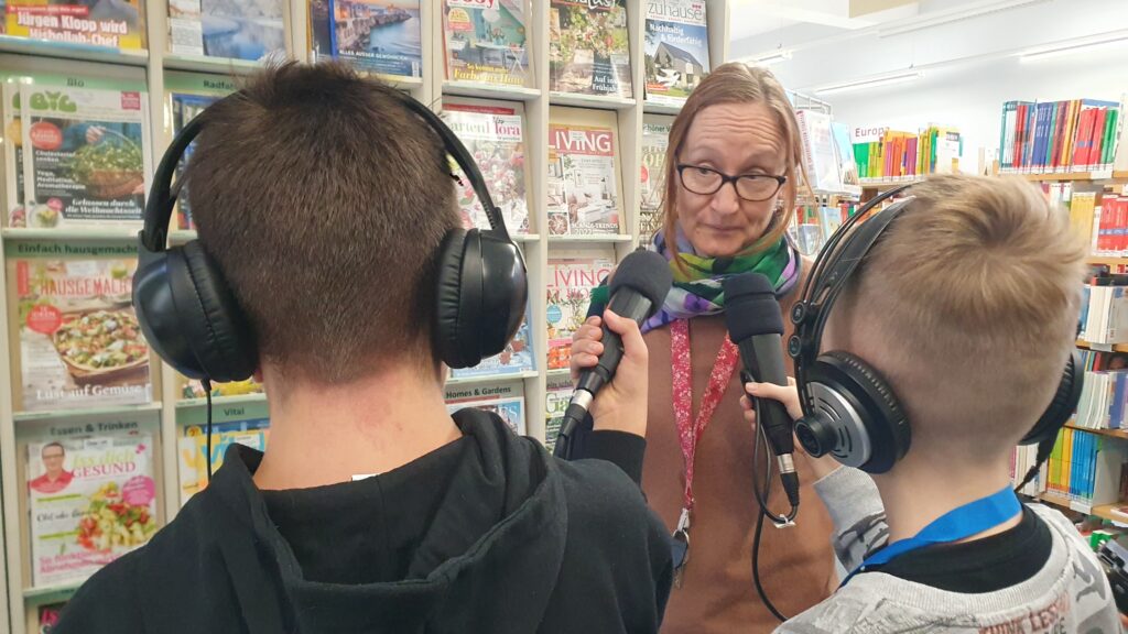 zwei Schüler mit Mikrofon und Kopfhörern interviewen Bibliothekarin vor einem Regal mir Reiseliteratur