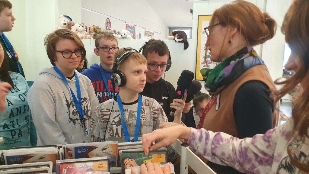 2 Schüler mit Mikrofon sprechen mit Bibliothekarin an einem Regal mit DVDs