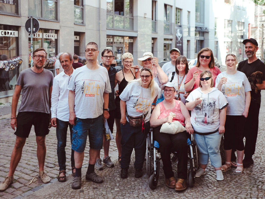 Eine bunte Gruppe von Menschen, darunter eine Frau im Rollstuhl, posiert und lächelt gemeinsam auf einer Kopfsteinpflasterstraße in einer städtischen Umgebung mit Geschäften und Schaufenstern im Hintergrund.