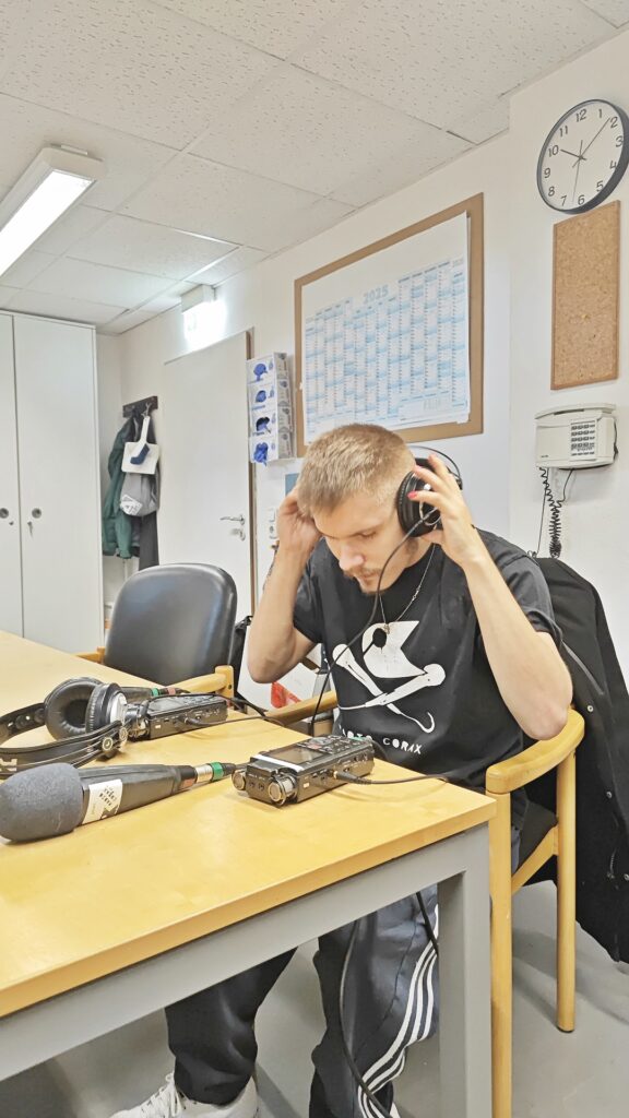 Mehrere Menschen sitzen und stehen an einem Tisch und unterhalten sich. Auf dem Tisch liegt ein Audio-Aufnahmegerät.