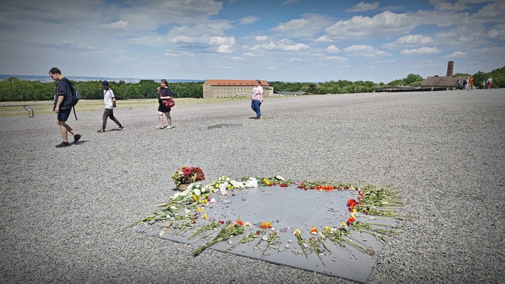 Eine quadratische in Kiesboden eingelassene Metallplatte erinnert an die Ermordeten des KZ Buchenwald. Auf den Rand der Platte wurden im Kreis angeordnet Blumen abgelegt und kleine Kieselsteine. Im Hintergrund ist das große Speichergebäude und der Schornstein des Krematoriums zu sehen.